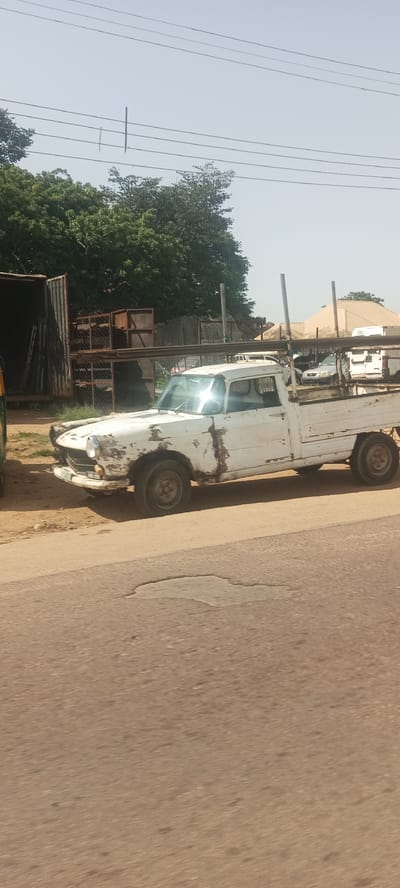 An aged white pickup truck, with visible rust and peeling paint, is parked on the side of a road in Rukuba Road, Nigeria. The truck has a long bed and appears to be carrying metal pipes vertically secured by poles. In the background, there are trees, some metal structures, and buildings with pitched roofs. The sky is clear and bright. The road in the foreground shows signs of wear, including a pothole.