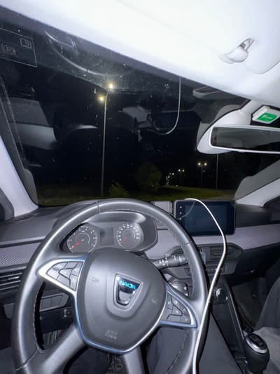 The image presents a nighttime view from within a Dacia passenger vehicle, specifically from the driver's perspective. The black steering wheel with the Dacia logo and integrated controls dominates the foreground. Behind it, the instrument cluster displays analog speedometer and tachometer dials, both indicating zero. A central touchscreen infotainment system is mounted on the dashboard. A white cable extends from the upper interior, across the infotainment screen, and descends toward the passenger footwell. A manual gear shift lever is visible to the right of the center console. Affixed to the upper windshield near the rearview mirror is a green Via Verde electronic toll transponder. Through the windshield, a dark outdoor environment is visible, illuminated by a series of amber streetlights lining a road or open area that recedes into the distance. Sparse vegetation is discernible beyond the immediate illuminated zone. The scene is located in Mirandela, Portugal.