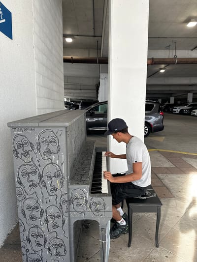 A young man in a grey t-shirt, black shorts, and a dark baseball cap is playing a piano in a parking garage. The piano is painted grey with black and white drawings of faces. A black bench is set up behind the piano for the player to sit. The parking garage has concrete walls and floors, with several cars parked in the background. The location is in the city of Tampa, United States of America.