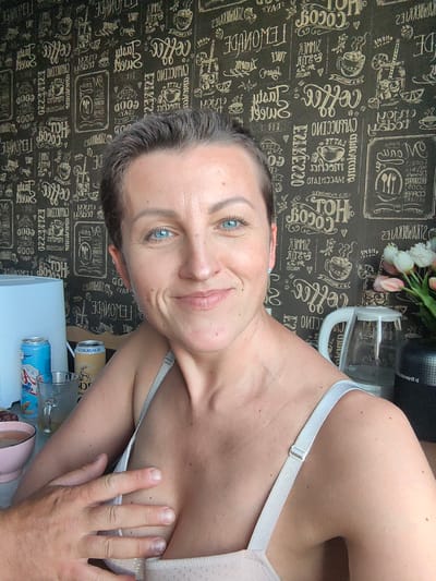 A woman with short, dark hair and blue eyes is pictured from the chest up, looking towards the camera. She is wearing a light-colored bra with visible straps. Her right hand is resting on her chest, partially covering her bra. The background is a dark wallpaper with a repeating pattern of text and illustrations related to coffee and drinks, featuring words like "Coffee," "Cappuccino," and "Lemonade."
To her left, a white humidifier or air purifier is visible, along with two cans of beverages and a glass of liquid. To her right, a transparent electric kettle is present, and behind it, a vase with pink and white tulips.
The lighting suggests it is daytime. The overall scene appears to be indoors, possibly a kitchen or living area, given the objects present. The woman's expression is a gentle smile.
Visible text on the wallpaper includes various phrases related to beverages and coffee shop themes, some of which are in English. There are also some Cyrillic characters visible within the patterns on the wallpaper.