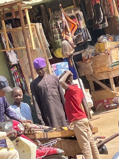 An outdoor market scene in Samaru, Nigeria, features five individuals gathered around a wooden stall under bright daylight. A central adult male, wearing a purple skullcap and a dark brown long-sleeved tunic, stands looking left with a slight smile. To his immediate left, a child in a white and light blue shirt also looks left. To the central male's right, a child in a red polo shirt and tan pants is seen from behind, with his right hand on the back of his head. In the lower left foreground, a person partially obscured by a dark cap is positioned on a red and white scooter/motorcycle, with hands on the handlebars. A wooden display platform in front of the central male and the child in white holds bundles of cut sugarcane. The background stall exhibits various packaged hair extensions, fabrics, stacked cardboard boxes, a hanging patterned scarf, and a light-colored wig form. The ground is dry earth.