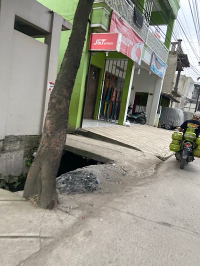 The image shows a street in Margaasih, Indonesia, at the coordinates -6.954 latitude and 107.551 longitude. A large tree trunk is located in the foreground and is growing at the edge of the street. The tree trunk is partially covering a large hole in the street, which is filled with dirt and debris. The road is paved with concrete and there is a small amount of debris and dirt scattered on the surface. There is a small building with a green facade and a sign that reads "J&T Express" to the left of the image. A person is riding a motorcycle with multiple green cylinders attached to the back. The person is wearing a black shirt and a white helmet. There is a small amount of pedestrian traffic in the background. The sky is overcast and there are power lines running across the street. The building has a small wooden door and a metal gate with colorful fabrics hanging inside the building. There is a small white cat visible on the sidewalk. A motorcycle with a rider is traveling away from the camera towards the left of the image. The motorcycle is carrying multiple green cylinders. The environment is urban and there are several buildings in the background. The sidewalk is cracked and uneven and there is a small drain in the foreground. The street is fairly narrow and the overall scene is one of a typical urban street in a developing country.