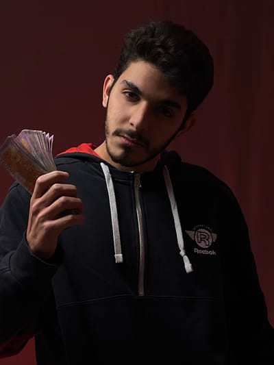 An image displays a young male, appearing to be in his late teens or early twenties, positioned against a solid, dark reddish-brown background. He has dark, short curly hair, a short beard, and a mustache. The subject is looking directly forward, making eye contact with the viewer. He is dressed in a dark, possibly black, hooded sweatshirt (hoodie) featuring a half-zip closure on the front and white drawstrings. The interior lining of the hood appears red. On the left chest area of the sweatshirt, a logo is visible, consisting of "INTERNATIONAL" text above a winged "R" symbol, with "Reebok" written beneath it. In his left hand, the individual holds a fanned stack of multiple banknotes, which appear to be Syrian Lira, with faint numbering suggestive of 2000 Lira denominations visible on some notes. The lighting illuminates primarily the subject's right side, creating shadows and contrast across his face and attire. The scene is located in Al-Mazza Municipality, Syria.