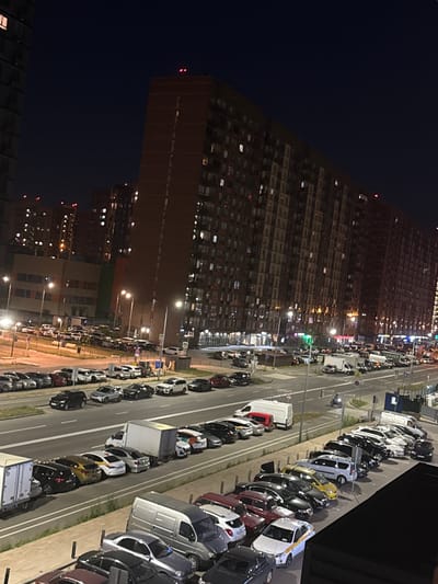 The image depicts a nighttime urban scene in Mirny, Russia, characterized by a large, multi-story residential building dominating the background. The building features numerous illuminated windows, suggesting occupied apartments. In the foreground and mid-ground, a street and parking areas are visible, lined with a variety of parked cars. Streetlights illuminate the scene, casting shadows and highlighting the parked vehicles. There are no visible people in the frame, and no apparent actions or interactions are taking place. The environment appears to be a typical residential area in a city, with the presence of vehicles and buildings indicating urban infrastructure.