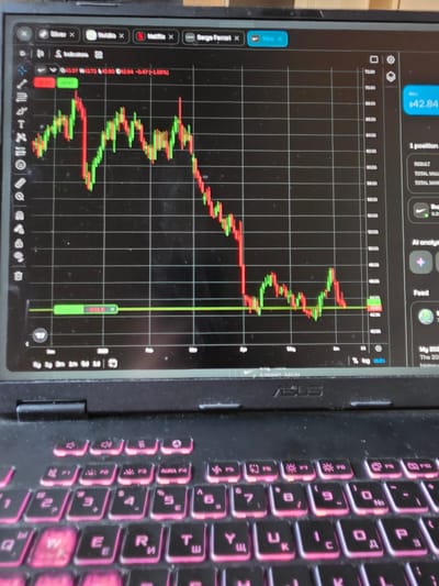 The image displays a close-up, slightly angled view of an open ASUS laptop, situated in the city of Dupnitsa, Bulgaria. The laptop's screen is illuminated, showcasing a financial trading platform with a dark interface. At the top of the screen, multiple tabs are open, including "Silver x," "Nvidia x," "Netflix x," "Serge Ferren x," and the currently active "Nike x," highlighted in blue.
The central part of the screen features a candlestick chart, illustrating price movements of Nike stock from approximately April to June. Green candlesticks denote periods of upward price movement, while red candlesticks indicate downward trends. A horizontal green line is present on the chart, marking a price level around 42.00-43.00, accompanied by a small green bar labeled "-0.01€." The vertical axis on the right side of the chart displays price increments ranging from 72.00 down to 40.00. To the right of the chart, a sidebar contains a "SELL" button indicating "€42.84," followed by details on "1 position," "RESULT," "TOTAL VALUE," "TOTAL MAR," a "Buy" section with a Nike logo, "AI analysis," and a "Feed" section. The bottom-left corner of the screen shows additional numerical data, "0.268887, $42.84."
Below the screen, the laptop's black keyboard is visible, featuring distinct pink backlighting on its keys. The keycaps display both Latin and Cyrillic characters. The function row includes specific keys for volume control, screen brightness, and an "AURA F4" key. The ASUS brand logo is embossed clearly on the laptop's body between the screen and the keyboard. The laptop rests on a dark, flat, and out-of-focus surface, suggesting an indoor environment. No individuals are depicted in the scene.
