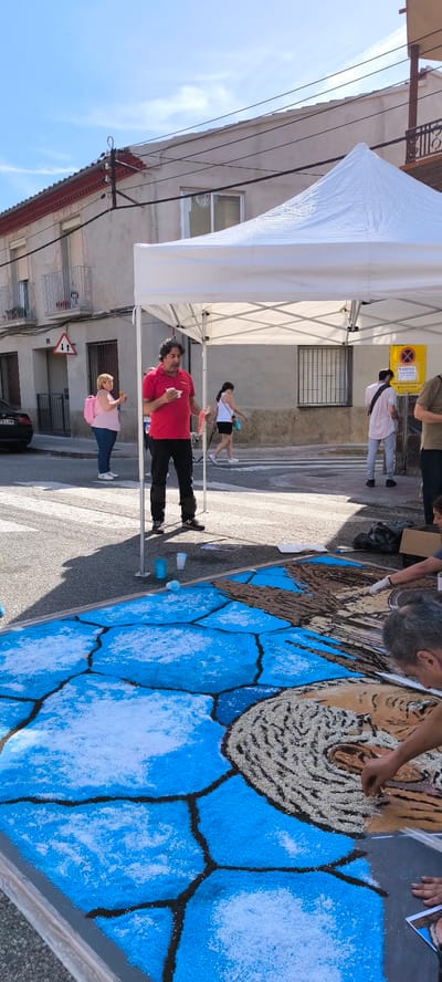 The image depicts an outdoor street scene in Ripollet, Spain, during daylight hours, focused on the creation of a large-scale ephemeral artwork on the asphalt pavement.

In the immediate foreground, a multi-paneled artwork is being assembled using granular materials. The left and lower sections consist of varying shades of blue, outlined in black, resembling a segmented pattern. The right and upper sections feature intricate patterns: one area is filled with light-colored, textured granular material, while an adjacent section is brownish-orange. Two individuals are actively engaged in placing granular materials onto the artwork. One person, wearing white gloves, meticulously applies light-colored grains to a patterned section. The hands of a second person are visible, also working on the patterned area. The artwork is partially shaded by a white pop-up canopy tent with visible metal framing.

Beneath the tent, centrally positioned, a man with dark, curly hair and a beard, wearing a red polo shirt and dark pants (with what appear to be knee pads or protectors), stands holding a white cup and a small white object. He is looking towards the left. On the asphalt near his feet, three plastic cups are visible: two blue (one upright, one overturned) and one white.

Behind the tent and the artwork, the street features a crosswalk with white stripes. To the left, a black car with a partially visible license plate "2478 LHW" is parked. A woman in a pink top and blue jeans, carrying a pink backpack, stands on the sidewalk, holding a light-colored food item. Above her, affixed to a building, is a white triangular traffic sign with a red border and two black arrows pointing vertically, indicating two-way traffic, with the number "24" below it.

To the right of the central standing man, a woman in a white tank top and dark shorts walks away from the camera, leading a small dog on a leash. Further right, a man in a light pink shirt and grey pants stands with his back partially to the camera. A yellow sign with a red 'X' and text, partially legible as "CORPUS," is posted on a building. Another individual in a brown t-shirt is partially visible on the far right.

The background consists of multi-story buildings with light-colored facades, windows, and some balconies with metal railings. Overhead, power lines traverse the scene against a bright blue sky with sparse white clouds.