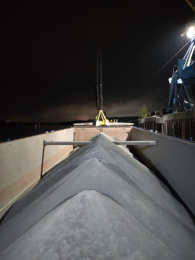 The image depicts the interior of a barge or ship filled with a gray, granular material, likely sand or gravel. A crane with a yellow grab bucket is positioned above the hold, presumably loading or unloading the material. The scene is illuminated by artificial lights at night. The barge is docked next to a pier or wharf, evidenced by the pilings and the structure to the right of the image. In the background, across the water, there is a hint of land with trees and distant lights, possibly indicating the city of Tver, Russia. The overall setting suggests a nighttime operation at a river or port facility.