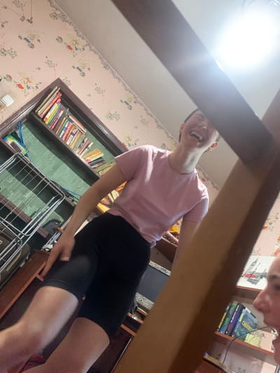 The image, taken in Hoholeve, Ukraine, shows a young person indoors, laughing with their mouth wide open. They are wearing a pink t-shirt and black shorts. To the left of the person is a wooden bookshelf filled with books. A drying rack is also visible near the bookshelf. The wallpaper in the background has a pattern of bunnies. A bright light source is present in the upper right corner, creating a glare. The overall scene suggests a casual, home environment.