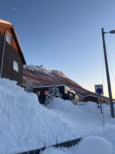 A snow-covered landscape features buildings and a mountain under a clear blue sky. The roof of a brown house with windows is visible in the foreground, while a dark building stands amidst deep snowdrifts. A mountain rises in the background. A streetlight and a "Sone" parking sign are placed above the buildings. A bright glow highlights the mountain's surface.