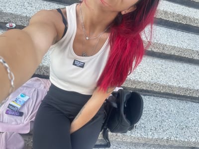 In Zheglartsi, Bulgaria, a person with vibrant red hair is seated on concrete stairs. They are wearing a white ribbed tank top with a black bra strap visible, black leggings, and a silver necklace. To their left, a pink jacket, a pack of "Omo" cigarettes, and a purple lighter are laid out. A black backpack is positioned to their right. The environment suggests an outdoor urban setting.