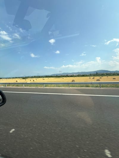 The image captures a view from a vehicle, looking towards a rural landscape under a bright blue sky with scattered clouds. In the foreground, a portion of a grey asphalt road is visible with a white line marking. To the left, the reflection of the car's interior and the sky is seen on the windshield, along with the side mirror. Beyond the road, a field of harvested crops, appearing as golden stubble, is dotted with numerous round hay bales. A metal guardrail separates the road from the field. In the background, a line of green hills stretches across the horizon, with some trees and power lines visible. The scene appears to be located in or near the village of Gorni Bogrov, Bulgaria, given the characteristic rural landscape and architecture implied by such locations. There are no people or animals directly visible in the frame. The time of day is daytime, likely late morning or afternoon, due to the quality of light.