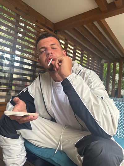 A man is captured in a mid-shot, appearing to be on a balcony or porch in Razlog, Bulgaria, during the daytime. He is oriented sideways, looking directly at the viewer. A lit cigarette is held between his lips, with his right hand positioned near his mouth. In his left hand, he holds a light-colored smartphone, which has a matching case.
The man is wearing a white t-shirt layered under a two-toned jacket. The jacket is primarily light-colored (beige or off-white) with black panels on the sleeves and sides, edged with white piping. The fabric has a subtle textured pattern, possibly a ripstop material. White drawstrings are visible at the bottom hem of the jacket.
The background consists of wooden architectural elements, including prominent vertical slats that form a railing or screen. Through these wooden structures, green foliage from trees or bushes is visible, suggesting an outdoor environment. To the man's right, a blue and white patterned cushion or upholstery indicates he is seated or reclining. The lighting suggests it is daytime. No visible text is present in the image.