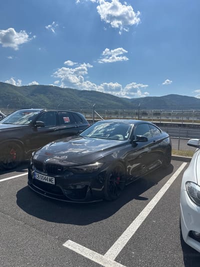 A black BMW coupe and a dark SUV are parked in a lot. The BMW has a license plate reading "CB 0044 ME". The SUV has a red sticker on its window. In the background, a fence with advertisements for "Monster" is visible, beyond which are hills and a blue sky with white clouds. The location is the race track in Dospey, Bulgaria.