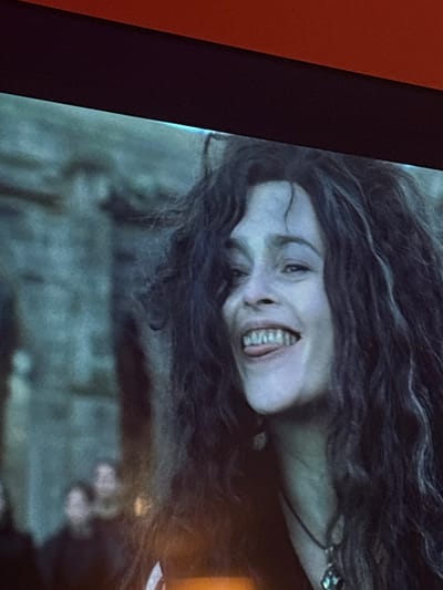 A screen displays a scene featuring a pale-skinned woman with dark, wavy hair framing her face. She has a sinister grin, exposing her teeth. The background is blurry, suggestive of a rugged, possibly outdoor, environment. The image seems to be captured from a television or monitor, as evidenced by the surrounding black frame. The lighting accentuates the woman's face, creating a dramatic effect.
