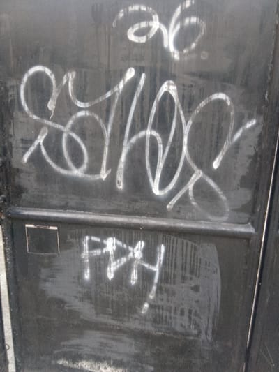 The image depicts graffiti on a dark, weathered metal surface, possibly a door or dumpster. The graffiti consists of two distinct tags. The upper tag is larger and more elaborate, rendered in white, and appears to be a stylized name or symbol. It features swirling lines, loops, and what may be the numbers "36" incorporated into the design. Below this, a smaller, cruder tag in white reads "FPHY" or something similar, with some surrounding smudging. The metal surface itself shows signs of age and wear, with scratches, scuffs, and a slightly uneven texture. The lighting suggests an outdoor environment, with a subtle sheen on the graffiti indicating recent application or a reflective surface. The image quality is somewhat grainy, typical of casual photography. The setting is identified as Cisarua, Indonesia, which implies this urban or semi-urban graffiti is located within that specific geographical area.