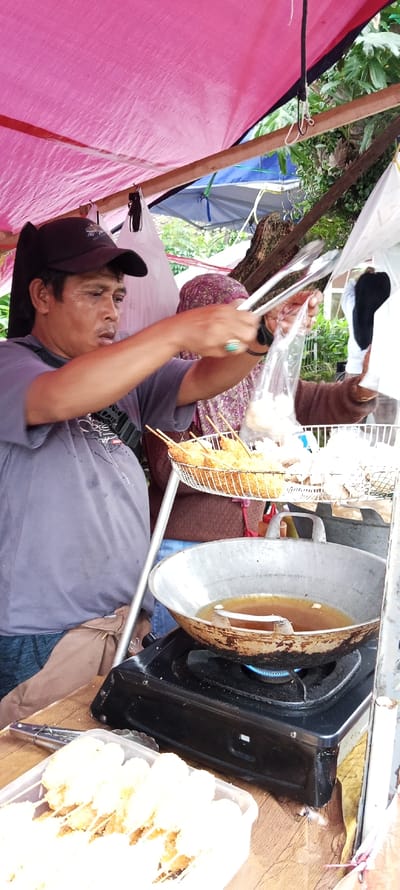 The image shows a food stall in the city of Banjaran, Indonesia, located at coordinates -7.046, 107.581. A man, wearing a grey shirt and a black cap, is standing behind a counter. He is using tongs to grab some food from a plastic bag held by another person, partially visible, who is wearing a brown sweater with a pink headscarf. The food is deep fried in a wok on a gas stove. Another plastic container is placed on the counter, containing more fried food. The food stall is covered by a pink tarpaulin. The background features a wooden structure with a green plant growing on it. The scene suggests that the man is operating the food stall and is preparing the food.