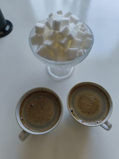 A top-down, medium shot captures two white coffee cups with dark, frothy coffee, positioned at the bottom of the frame on a white surface. To the upper center, a glass bowl filled with sugar cubes is visible. The scene is evenly lit, creating a clean and bright atmosphere. The composition is balanced, with the sugar bowl and coffee cups occupying distinct areas of the frame. The surfaces appear clean and uncluttered, suggesting a well-maintained setting. There are no people or animals present in the image. The environment is sterile and appears to be indoors. This image likely represents a moment of hospitality or a simple coffee break.