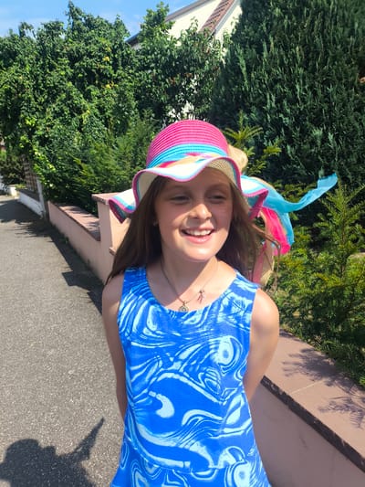 A young girl with long brown hair smiles brightly as she stands outside on a sunny day. She is wearing a colorful wide-brimmed hat with pink, blue, and yellow stripes, and a blue dress with a white swirling pattern. A delicate necklace with a small pendant rests on her collarbone. The girl is positioned in front of lush green trees and a light-colored wall. To her left, a paved pathway is visible, with a shadow of what appears to be the person taking the photo cast on it. The setting suggests a warm, pleasant day, likely in the afternoon, given the strong shadows. The overall mood is cheerful and lighthearted. The location is identified as Ittenheim, France.