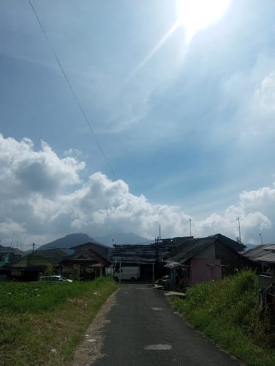 The image displays a daytime scene in Đăk Pxi, Vietnam, featuring a narrow, dark grey paved road extending from the foreground into the middle ground. The road is bordered by unkempt green grass and foliage. On the left, multiple buildings with dark roofs are partially visible behind vegetation, with a light-colored vehicle obscured further back. On the right, a prominent building features a pinkish facade and a dark roof, adjacent to a smaller, darker structure. At the termination of the visible road, a white van is parked beneath a covered area with an unreadable banner. Utility poles and wires are distributed among the structures. The background reveals green, forested mountains beneath a blue sky populated by scattered cumulus clouds. A strong sun flare originates from the upper right, and a thin, dark wire extends diagonally across the upper left sky. A small red and white flag is visible on a pole above the buildings against the mountain backdrop. No individuals are depicted.