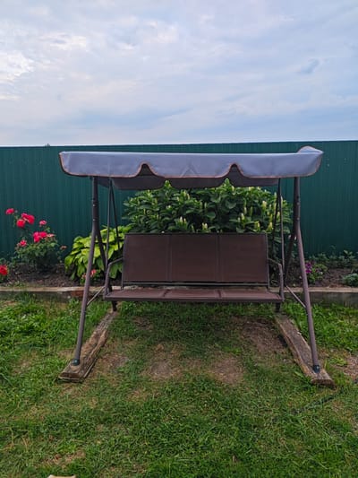A garden swing is positioned on a grassy lawn in front of a dark green corrugated metal fence. The swing has a dark brown metal frame and a brown fabric seat and backrest. A grey canopy with a scalloped edge provides shade. Behind the swing, a bed of green plants with white flower spikes is visible, along with some red roses to the left. The ground around the swing is covered in green grass, with two weathered wooden planks placed under the swing's legs for stability. The sky above is cloudy. The image appears to have been taken outdoors in a residential area.
