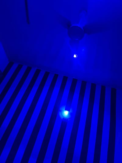 The image captures a dimly lit interior space, dominated by vibrant blue lighting. On the left side, a ceiling fan with white blades is visible, casting a shadow on the blue wall. Adjacent to the fan, a small, bright light fixture emits a white glow.

The right side of the image features a wall with a distinctive pattern of alternating dark blue and black horizontal stripes. Another bright, white light fixture is positioned on this striped wall, illuminating a section of the stripes and creating a glare. The overall impression is one of a modern, possibly decorative or functional, lighting setup in a room. There are no people or any visible text in the image. The lighting creates a dramatic and somewhat abstract atmosphere. The setting appears to be indoors, with the blue light suggesting either specialized lighting or a filter effect. Given the provided location context of Pantinapu, Nigeria, the scene depicts an interior environment within that region.