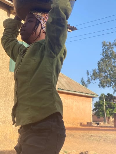 An outdoor scene in Udu, Nigeria, depicts a young individual in profile, facing left, carrying a large, dark basin on their head, hands raised for support. The individual wears an olive green long-sleeved collared shirt, dark trousers, and a patterned headwrap over braided hair. The immediate left foreground shows a light brown building wall with a partially visible green shutter. In the midground, under a clear blue sky with overhead utility wires, an orange-walled building with a corrugated metal roof stands beside a dusty, reddish-brown unpaved area. Further back, additional buildings and trees with sparse foliage are visible. A dark-colored animal, consistent with a dog, walks in the distant background on the unpaved ground. Sunlight illuminates the scene, indicating daytime conditions.