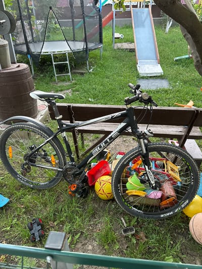 A black and white mountain bicycle is leaning against a wooden bench in a grassy backyard. The bicycle has the brand name "STYX" visible on its tire and "ACE OF PACE" written on the side of the tire. Various colorful toys, including a red plastic bin, a yellow ball, a pink object, and green and yellow toy pieces, are scattered around the bicycle and the bench. A basketball and a large yellow ball are also present in the scene. In the background, a trampoline with a black net is visible, as well as a wooden barrel. The setting appears to be outdoors during the day, with the sun casting shadows. The overall impression is one of a cluttered but lively outdoor play area. Text on the bike includes "STYX", "ACE OF PACE", and "SUNTOUR". The location is Pîrlița, Moldova.