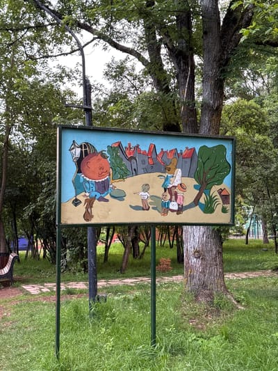 The image shows a painted sign depicting a whimsical scene with stylized figures. The sign is rectangular and appears to be made of wood, framed by dark green metal. The artwork features a cartoonish style, with bright colors and simplified forms.

In the foreground of the painting, a large, round-headed figure with an orange face and rosy cheeks is depicted. This figure is wearing a blue coat and a dark hat, and is holding what appears to be a book. To its left, a vintage-style lantern is placed on the ground. Further back, a group of smaller figures are gathered. One figure, wearing a pink hat and a white dress, is leaning over, possibly interacting with the others. Another figure wears a blue hat and a white shirt. They are all positioned in front of a row of stylized red-roofed buildings.

Behind the figures and buildings, a large green tree is prominently featured, its branches reaching upwards. The background of the painting includes a light yellow sky and hints of other structures. The overall scene suggests a playful, perhaps educational, depiction of community life or a story.

The sign is mounted on a dark metal pole and is surrounded by lush green grass and mature trees, suggesting an outdoor setting like a park or a public garden. The thick trunks of the trees and the dense foliage indicate a natural environment. The ground around the sign is partly covered by a stone pathway. The time of day appears to be daytime, with diffused light suggesting an overcast or shaded condition. The location context is Dilijan, Armenia. There is no visible text in the image.