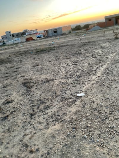 The image depicts an unpaved, uneven plot of land in الغنادة, Tunisia, under a sunrise or sunset sky. The foreground and midground consist of a dirt and gravel surface, exhibiting tire tracks and scattered refuse, including blue plastic items and a crumpled white object. In the midground, several structures are visible, appearing to be residential or commercial buildings under construction, constructed from concrete blocks and reddish-brown brick. Some buildings are multi-storied and incomplete, with exposed walls and rooflines. Piles of construction materials, specifically what appears to be gravel or sand, are present near some of these structures. A white van or truck is parked near the buildings on the left side of the frame. The horizon is marked by a line of dark vegetation, beyond which the sky displays a prominent gradient of orange and yellow hues, indicative of dawn or dusk, with sparse, light cloud formations. No individuals or active interactions are visible within the scene.