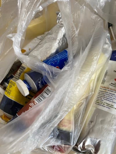 The image shows a close-up, overhead view of several tubes of paint contained within a clear plastic bag. The tubes are of various colors and brands, with some appearing to be oil paints based on the labels and metallic foil packaging. One tube is yellow with "120 ml 4 US FL. OZ." printed on it, and a blue cap. Another tube is dark blue with a silver cap, and some blue paint is squeezed out around the cap. A brown tube is also visible, with a barcode. Some of the tubes have labels with text in what appears to be Czech. A label on one tube reads "Umelecká akrylová" and another section states "VAROVÁNÍ: Odstraňte obal..." which translates to "WARNING: Remove packaging...". The background is out of focus but suggests an indoor setting with neutral lighting. There are no people visible in the image, and no specific location in the city of Bílina, Czechia, can be identified from the visual information provided.
