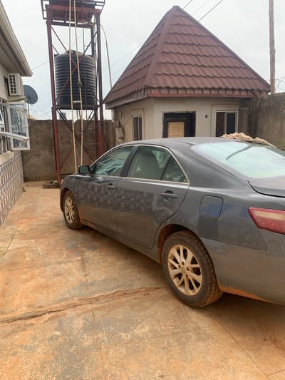 In Kakau, Nigeria, a gray sedan is parked on a dirt-covered concrete driveway in front of a building. The car is viewed from the rear passenger side, showing its right side and rear wheel. The vehicle appears dusty, with dirt splattered on the lower body panels and wheel. A light-colored cloth is draped over the rear windshield.
Behind the car, a tall, dark water tank stands on a metal scaffolding, with pipes extending from it. To the right of the tank is a building with a brown, tiled, pitched roof and a lighter colored facade. A concrete wall, topped with barbed wire, runs along the right side of the property. To the left of the water tank and partially visible is another building with a tiled wall and what appears to be an air conditioning unit mounted on its exterior. The ground is a mix of concrete and reddish-brown dirt. The sky is overcast.