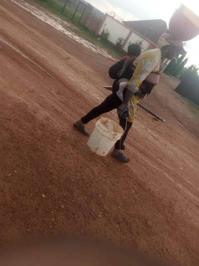 A low-angle, slightly tilted, medium shot captures a person carrying a child on their back and a bucket on the ground. The person, facing right and angled away from the camera, wears a yellow and white patterned blouse and black pants, with gray clogs on their feet. They are carrying a light-colored bucket in their right hand. A child is secured to their back with a cloth wrap. On the person's head, a pink and yellow bowl-shaped object is balanced. The ground is composed of reddish-brown dirt, with patches of water indicating recent rain. In the background, a white wall with a fence is visible, along with trees and a structure. The sky is overcast. The scene appears to be outdoors, on a dirt road or path, likely in a rural or semi-urban setting, potentially in the city of Kwang, Nigeria, given the soil color and vegetation.
