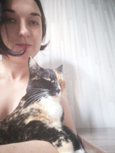 A woman with dark hair, seen from the chest up, is holding a calico cat. The woman's face is turned towards the camera, with her eyes partially closed and a slight smile. She is wearing a white top, with her shoulders exposed. The cat is looking towards the right of the frame, with one eye closed and its head tilted. The cat's fur is a mix of black, orange, and white. The background appears to be a wall with vertical lines, possibly a curtain or a textured wallpaper. The lighting is soft and diffused, suggesting an indoor setting. The overall impression is a casual and intimate portrait of a woman and her pet. The image was taken in Tosno, Russia.