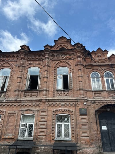 The image shows the facade of a red brick building, likely an old historical structure, with an ornate architectural style. The building has multiple windows, some of which are open, and decorative brickwork. Above the windows, there are elaborate brick cornices and pediments. The sky above is blue with scattered white clouds. A dark wire is visible running across the top of the frame. On the right side of the building, there is a large dark entrance door and a sign with text. There are no people visible in the image. The overall impression is of a weathered but historically significant building.