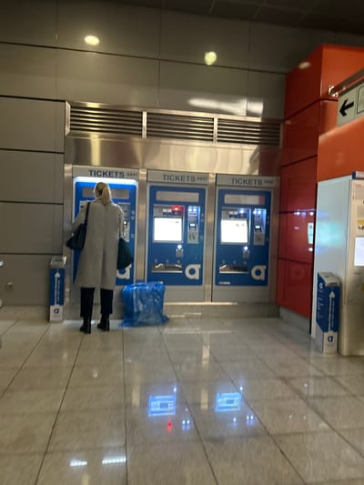 The image captures an indoor scene at a modern public transport station, consistent with the Koridallos Metro Station in Greece. The viewpoint is slightly angled, looking towards a row of ticket vending machines.
In the foreground, a woman with blonde hair, wearing a light-colored long coat, black pants, and black boots, stands with her back to the camera. She is actively interacting with one of the blue and silver ticket vending machines, appearing to purchase a ticket. A dark handbag is carried on her shoulder.
There are several identical ticket machines arranged in a line, each labeled "TICKETS" followed by an alphanumeric identifier such as "A048", "A047", and "A041". These machines feature screens, card readers, and slots for tickets or money. A distinct "b" logo of the public transport operator is visible on the side of each machine. A red indicator light is illuminated on the interface of one machine. A crumpled blue plastic bag lies on the floor next to the middle machine. Near the woman's feet, a small blue and white box, possibly a waste bin or information dispenser, shows an arrow and partially visible text that includes a letter "D".
The station environment is clean and modern, with light-colored tiled walls and a highly polished, light-colored tiled floor. The floor prominently reflects the bright screens of the ticket machines and overhead lighting, creating distinct glowing patterns. Above the machines, a reddish-orange architectural element is present, featuring a sign with a downward-pointing arrow. To the left, a vertical metal grate, likely for ventilation, is integrated into the wall. The scene suggests a functional and orderly public space.