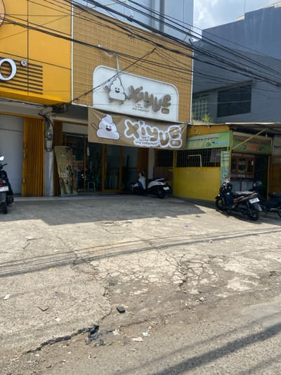The image shows a storefront with a sign that reads "XiYue Ice cream and happiness" in English and Chinese. The storefront is painted yellow and has a large, cracked, gray concrete area in front of it. Two motorcycles are parked in front of the store, one on the left side and one on the right side. The motorcycle on the left side is black with a yellow license plate, and the motorcycle on the right side is black with a white license plate. There are also a few small pieces of trash on the ground. The building next to the storefront is also painted yellow and has a sign that reads "Mie Ayam KUDAL M. Ihsan" in Indonesian. There is a small, open-air food stand next to the storefront on the right, with a sign that reads "BASO MALANG KUDAL SININ".  The image was taken in front of a storefront located in Kecamatan Margahayu, Indonesia, at the coordinates -6.963 latitude and 107.578 longitude.