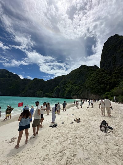 The image captures a bustling beach scene at Maya Bay, located within the Phi Phi Islands, near Ao Nang, Thailand. The foreground is dominated by a white sandy beach with a scattering of flip-flops and bags. Several groups of people are present, mostly tourists, dressed in casual beachwear and some in more formal attire, suggesting a range of activities. Some are walking along the shore, while others are standing in small groups, conversing or observing the surroundings. The turquoise waters of the Andaman Sea gently lap the shore, and a red flag is planted on the beach, possibly indicating a warning or a designated area. Towering limestone karsts covered in lush green vegetation form a dramatic backdrop to the bay, characteristic of the Phra Nang Cave Beach area. The sky is a mix of bright blue and scattered white clouds, with a subtle rainbow visible in the upper left quadrant. The overall atmosphere is one of a popular tourist destination experiencing a day with varied weather conditions.