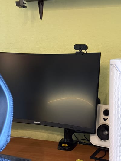 A curved Samsung monitor is positioned on a wooden desk. A webcam is mounted on top of the monitor. To the right of the monitor, a white speaker is visible. The wall behind the desk is painted a light yellow color. The scene appears to be an indoor setting, likely a home office or gaming setup. There is no indication of the city of Irpin, Ukraine in the image.