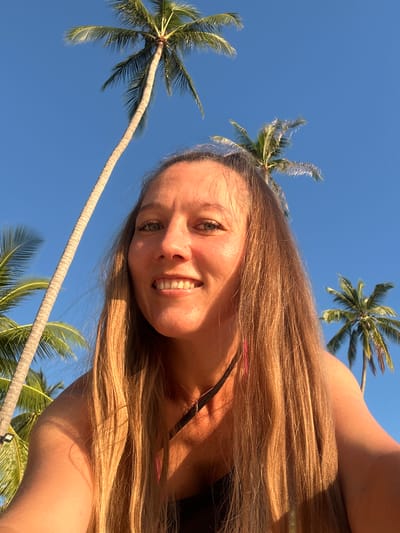 A woman with long, blonde hair smiles for a selfie outdoors under a clear blue sky, with several tall palm trees visible in the background. The setting appears to be a tropical location, possibly Baan Taling Ngam, Thailand, given the presence of palm trees and bright sunlight. The woman is looking directly at the camera, and her face is illuminated by the sun. She is wearing a black top with a thin strap visible.