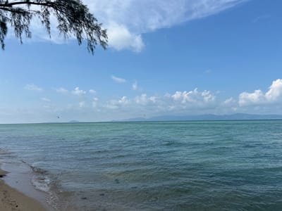 The image captures a coastal scene in Moo 1, Thailand, featuring a beach with light-colored sand, calm turquoise water, and a clear blue sky with scattered clouds. A tree branch with dark foliage is visible in the upper left corner. In the distance, a faint mountain range or islands are apparent. Two small kites or parasails are visible over the water. The water surface shows gentle ripples and slight variations in color due to depth or underwater features.