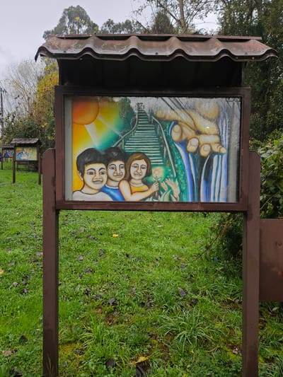 The image shows a series of wooden signposts in a park or outdoor area, likely in Osorno, Chile. Each signpost has a small roof and a framed picture or illustration behind glass. The visible artwork depicts a family of three with a hand reaching down from above. The signposts are spaced apart and situated on a patch of green grass. The weather appears overcast or wet.