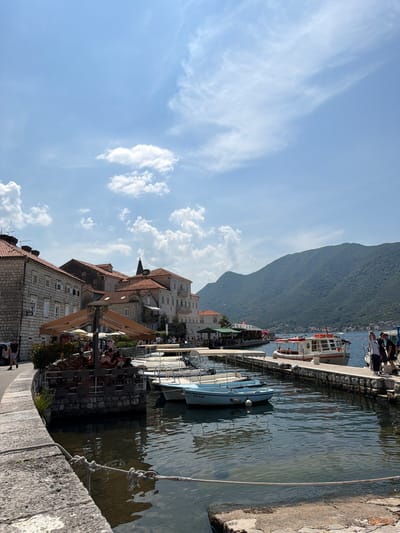 The image depicts a sunny day scene in Perast, Montenegro, with a clear blue sky and scattered white clouds. The foreground shows a stone waterfront with a thick rope tied to cleats, leading to the water where several small boats are moored. Further out, a larger passenger boat with a red stripe is docked at a pier, alongside other vessels. A row of stone buildings with terracotta roofs lines the waterfront, some featuring outdoor seating with umbrellas, suggesting a cafe or restaurant area. People are visible walking along the waterfront and seated at the outdoor tables. In the background, lush green mountains rise dramatically from the water's edge, framing the picturesque coastal town. The water is calm and reflects the sky and surrounding structures.