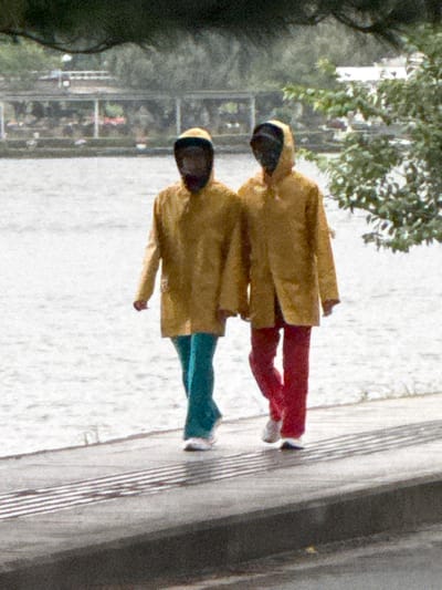 Two figures, dressed in hooded yellow raincoats and dark pants, walk side-by-side along a paved pathway adjacent to a body of water, likely Xuân Hương Lake in Đà Lạt, Vietnam. The figure on the left wears turquoise pants and white sneakers. The figure on the right wears red pants and white sneakers. The water is calm and reflects the overcast sky. Trees and distant structures, possibly part of the city's landscape, are visible in the background. The overall atmosphere suggests a cool or rainy day, as indicated by the raincoats.