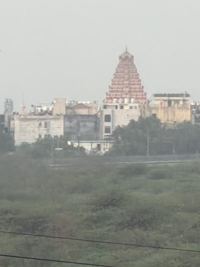 The image captures a distant view of a temple with a distinctive tiered, pyramidal roof, likely a Shikara, characteristic of Indian temple architecture. Adjacent to the temple are several buildings of varying heights and architectural styles, suggesting an urban or semi-urban setting. The structures appear to be a mix of older, possibly traditional buildings and more modern constructions. A significant portion of the foreground is occupied by lush green vegetation, possibly a park or undeveloped land, with several thin, dark horizontal lines that could be power lines or fences. The sky is overcast and hazy, giving the scene a muted, diffused light. The overall impression is one of a developed area with a prominent religious structure amidst other urban buildings and green spaces, under a cloudy sky. Without further context or identifying markers, pinpointing the exact location within Delhi is not possible from this image alone.