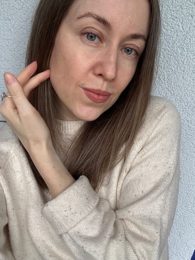 A Caucasian woman with light brown hair and blue eyes is pictured in a close-up shot. She wears a beige, speckled knit sweater. Her left hand is near her face, and a ring is visible on her finger. The background is a light-colored textured surface. There's no specific information to determine the location in Kudrovo, Russia.