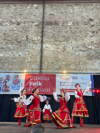 This media file captures a vibrant folk festival performance in Calella, Spain. The primary subjects are several young women dressed in traditional, richly embroidered red and white folk costumes. The costumes feature intricate floral patterns, wide red skirts with gold trim, white blouses with voluminous sleeves, and red leather boots. Some of the performers are also adorned with floral headpieces.
The scene takes place outdoors on a stage with a dark backdrop. To the left, a weathered stone wall serves as a textured background. A large, red banner is prominently displayed behind the performers, featuring white text that reads "Calella Folk Festival." Additional text on the banner includes "Ajuntament de Calella" (Town Hall of Calella), "FORTUNI VIATGES," and social media handles like "+ Folklore Festival Association," "folklore_festiva," and "y.folkloret".
The performers are engaged in dynamic folk dancing, their poses suggesting movement and energy. Their facial expressions convey joy and concentration as they execute their performance. The lighting suggests it might be late afternoon or early evening, with a warm glow visible on the banner. The overall atmosphere is festive and celebratory, highlighting cultural heritage through traditional dance and attire.