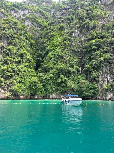 A white boat with a blue canopy is anchored in a bay of vibrant turquoise water. The boat has "PHRA NANG 2" written on its side. The water is calm and clear, with a line of yellow buoys stretching across the frame. In the background, towering limestone cliffs covered in lush green vegetation rise dramatically from the water. A small opening or cave is visible in the cliff face. A few people are visible in the water near the buoys, some appear to be swimming or snorkeling. The scene is idyllic and appears to be a popular tourist destination.
