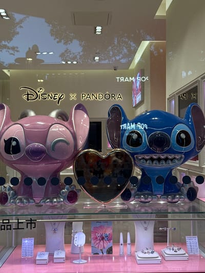 The image depicts a retail window display featuring Disney and Pandora collaboration merchandise, specifically two large character statues, one pink (Angel) and one blue (Stitch), positioned in the foreground. Between the statues, a metallic heart-shaped object is visible, inscribed with text. Both statues are large, detailed, and appear to be made of a glossy material. They are placed on a reflective surface that mirrors the ceiling lights and shadows of trees. Below the statues, a glass shelf displays various jewelry items, including rings, bracelets, and necklaces, arranged on white stands and boxes. A pink card with an image of Stitch and Angel is also displayed. The background shows store signage, including a large "Disney x Pandora" logo, and further display cases. The scene suggests a brightly lit retail environment. The text "品上市" is visible on the lower left side of the image, which translates to "product launch" or "new arrival" in Chinese. While the image was taken in Pudong, China, this specific location detail cannot be inferred solely from the visual elements.