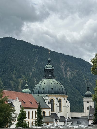The image shows a view of Ettal Abbey in Ettal, Germany, a Benedictine monastery at the foot of the Ettal mountain. The scene is dominated by the abbey's white Baroque architecture, featuring prominent green domes and a tall steeple. In the background, a densely forested mountain rises, partially obscured by a cloudy, overcast sky. The foreground shows the red-tiled roofs of some of the abbey's buildings and a stone wall. No people are visible in the image. The overall atmosphere is solemn and picturesque, with the architecture of the abbey standing in stark contrast to the natural beauty of the mountainous landscape.