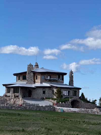 The image displays a large, multi-story stone and wood building with a complex roofline, situated on a grassy hill under a bright blue sky with scattered white clouds. The building features numerous stone chimneys, prominent stone foundations, and a mix of stone and wooden exterior walls. Large windows are visible on various levels. The surrounding environment includes more stone landscaping, a gravel path leading to an arched entrance, and some sparse vegetation, including trees on the right side. The foreground is dominated by a green, somewhat dry grassy field. There are no people or animals visible in the scene. This building is located in Gela, Bulgaria.